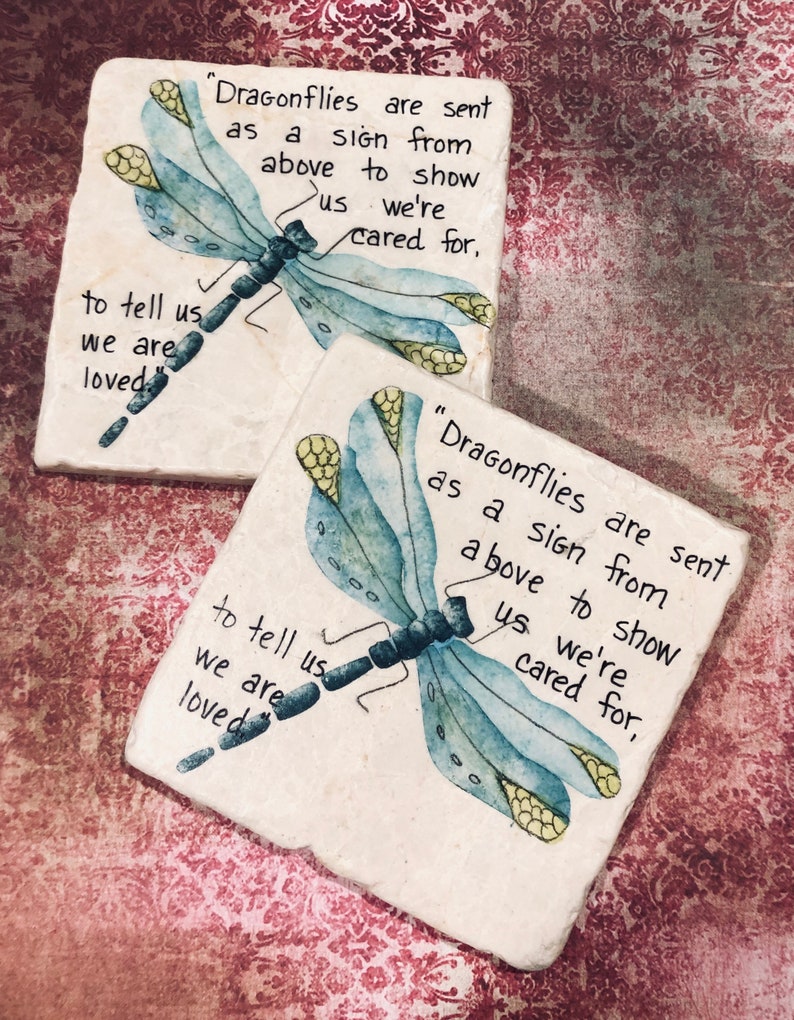 May include: Two white square coasters with a blue dragonfly painted on each. The dragonfly has yellow accents. The text on the coasters reads "Dragonflies are sent as a sign from above to show us we're cared for, to tell us we are loved".