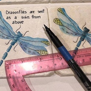 May include: Two white ceramic coasters with blue dragonfly illustrations and the text "Dragonflies are sent as a sign from above". A pink protractor and a black marker are on top of the coasters.