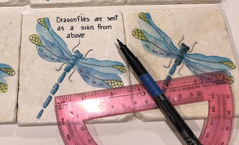May include: Two white ceramic coasters with blue dragonfly illustrations and the text "Dragonflies are sent as a sign from above". A pink protractor and a black marker are on top of the coasters.
