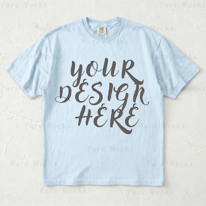 Può includere: T-shirt azzurra con la scritta "YOUR DESIGN HERE" in corsivo grigio scuro. La maglietta ha maniche corte e girocollo. Il tessuto sembra morbido e confortevole, adatto all'abbigliamento casual.
