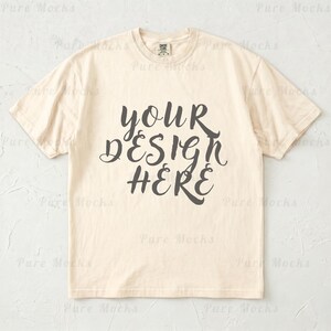 May include: Cream-colored t-shirt with the text "YOUR DESIGN HERE" in a dark gray, cursive font. The shirt has a classic crew neck and short sleeves. The shirt is a neutral color, suitable for various design applications.