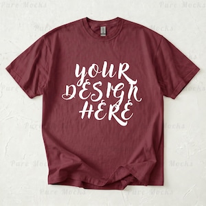 以下が含まれることがあります： 白文字で「YOUR DESIGN HERE」と書かれた、マルーン色のTシャツ。半袖とクルーネックです。首元にはGildanのブランドタグが見えます。ニュートラルな背景に表示されています。