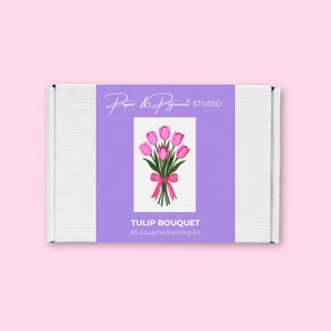 Puede incluir: Una caja blanca con una etiqueta morada que presenta una ilustración de acuarela de un ramo de tulipanes rosas atado con una cinta rosa. El texto de la etiqueta dice "Paper & Pigment Studio" y "TULIP BOUQUET A5 Gouache Painting Kit."
