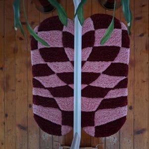 May include: Two matching, oval-shaped rugs with a checkerboard pattern in shades of pink and burgundy. The rugs are made of a plush, textured material. The background is a wooden wall.