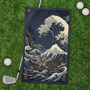 May include: A navy blue golf towel featuring a Japanese woodblock print of a large ocean wave with a boat and a person. The towel has a dark blue border and a metal grommet for hanging. Two golf balls and a golf club are on the green grass.