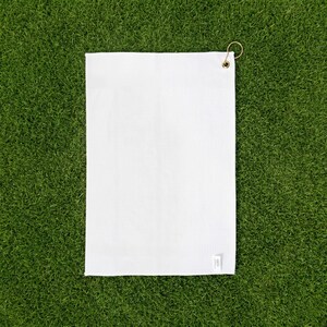 May include: A rectangular white waffle-weave towel featuring a small brass-colored metal grommet in the top right corner. The fabric has a distinct grid-like texture throughout, and a small white fabric tag is attached to the bottom right edge.