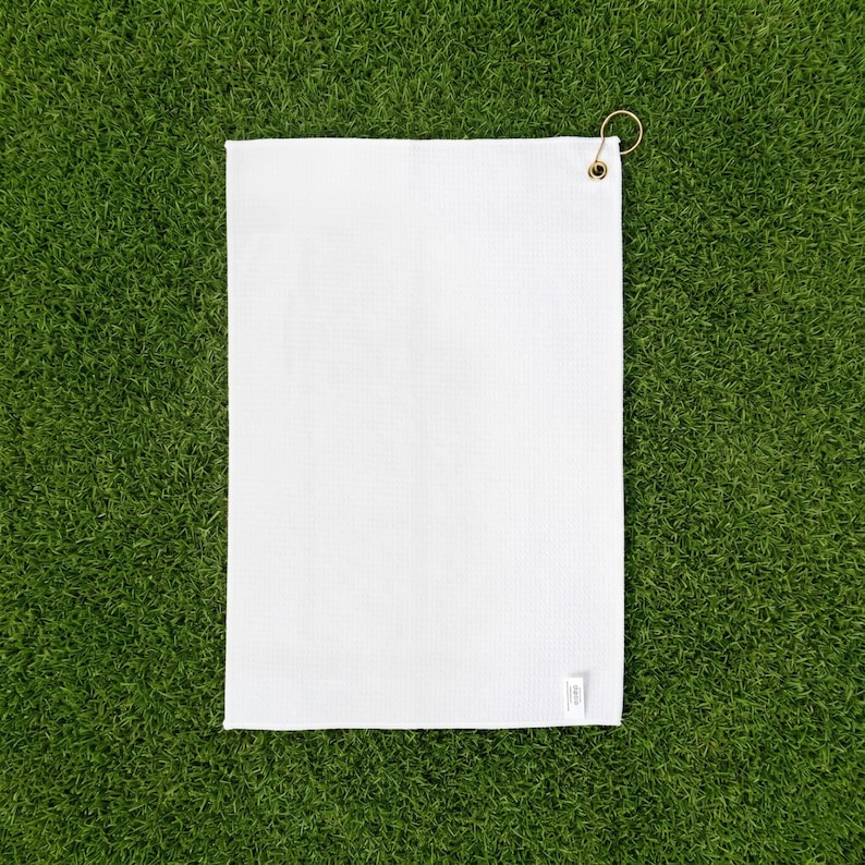 May include: A rectangular white waffle-weave towel featuring a small brass-colored metal grommet in the top right corner. The fabric has a distinct grid-like texture throughout, and a small white fabric tag is attached to the bottom right edge.