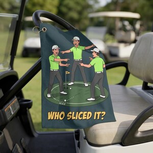 May include: A dark teal golf towel featuring a cartoon illustration of three golfers in lime green shirts and grey pants pointing at each other on a putting green. The text "WHO SLICED IT?" is printed in yellow at the bottom of the towel.