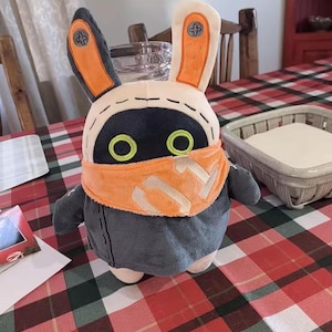 May include: A plush toy featuring a dark gray body, long ears with orange accents and screw-like details, and bright green circular eyes. The character wears an orange neck scarf with a light-colored graphic design and has visible stitching.