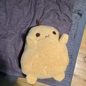 May include: A plush toy shaped like a rounded, tan potato with a fuzzy texture. It features two small, dark brown circular eyes and a simple, stitched horizontal line for a mouth. A small brown stem-like detail is visible at the top center.