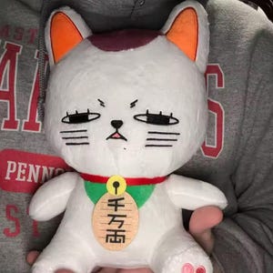 May include: A white plush cat with orange-tipped ears, a red collar, and a green bib. The cat has a grumpy expression with black eyes and whiskers. A wooden tag with Japanese characters hangs from the collar. The cat's paws have pink pads.