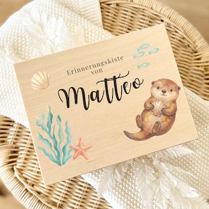 Könnte beinhalten: Eine Holz-Andenkenbox mit dem Namen "Matteo" in Schreibschrift. Die Box zeigt Aquarellillustrationen eines Otters, Muscheln, Seetang, eines Seesterns und kleiner blauer Fische. Der Text "Erinnerungskiste von" ist ebenfalls vorhanden.