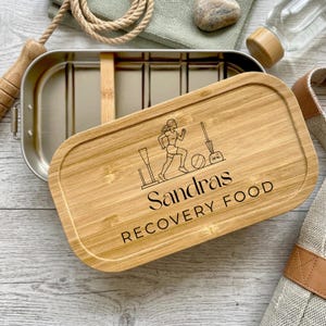 Könnte beinhalten: Eine rechteckige Lunchbox aus Edelstahl mit abnehmbarem Bambusdeckel. Der Deckel zeigt eine Lasergravur einer laufenden Person, eines Balls und einer Kettlebell sowie den in Schwarz gedruckten Text "Sandras RECOVERY FOOD".