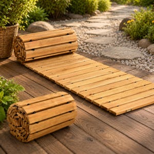Puede incluir: Alfombra enrollable de exterior de madera, hecha de listones de color marrón claro. La alfombra está parcialmente enrollada, con una sección completamente extendida. También se ven dos alfombras enrolladas adicionales. Las alfombras están sobre una terraza de madera, con un camino de piedra y vegetación en el fondo.