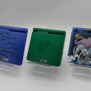Puede incluir: Tres consolas Nintendo Game Boy Advance SP. Una es azul con un diseño estilizado, otra verde con una silueta de personaje y la tercera azul con un gráfico de Pokémon. Todas están sobre soportes transparentes.
