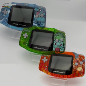 May include: Three custom Game Boy Advance consoles. The top console is blue with a Pokemon design and a Totodile graphic. The middle console is green with a Bulbasaur graphic. The bottom console is orange with a Charizard graphic.