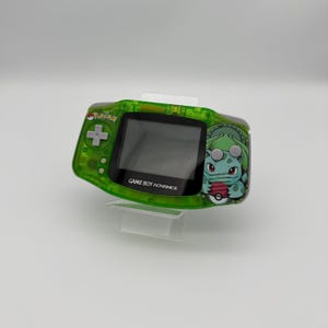 May include: A green Game Boy Advance handheld console with a Bulbasaur graphic on the right side. The console has a directional pad, buttons, and a screen. The text "GAME BOY ADVANCE" is printed below the screen. The console is on a clear stand.