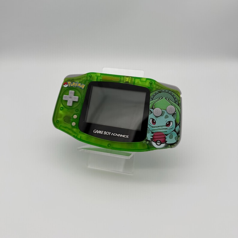 May include: A green Game Boy Advance handheld console with a Bulbasaur graphic on the right side. The console has a directional pad, buttons, and a screen. The text "GAME BOY ADVANCE" is printed below the screen. The console is on a clear stand.