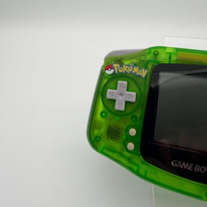May include: A transparent green Game Boy Advance handheld console. The console features a directional pad, buttons, and the word "Pokemon" above the directional pad. The screen is black, and the words "Game Boy" are visible on the right side.
