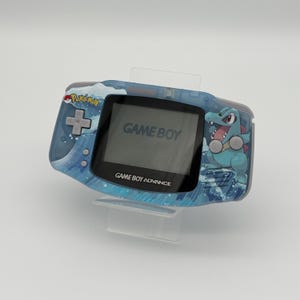 May include: A blue Game Boy Advance handheld game console with a Pokemon design. The console features a screen displaying "GAME BOY" and "GAME BOY ADVANCE". A cartoon crocodile is on the right side of the console.