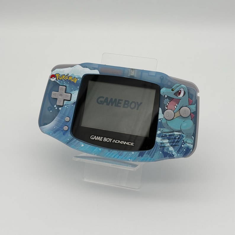 May include: A blue Game Boy Advance handheld game console with a Pokemon design. The console features a screen displaying "GAME BOY" and "GAME BOY ADVANCE". A cartoon crocodile is on the right side of the console.