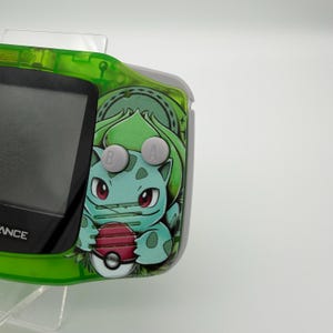 May include: A green and gray handheld gaming device with a Bulbasaur graphic. The device has a screen, gray buttons, and a green frame. The Bulbasaur graphic is in shades of green, blue, and red, holding a Pok&eacute;ball.