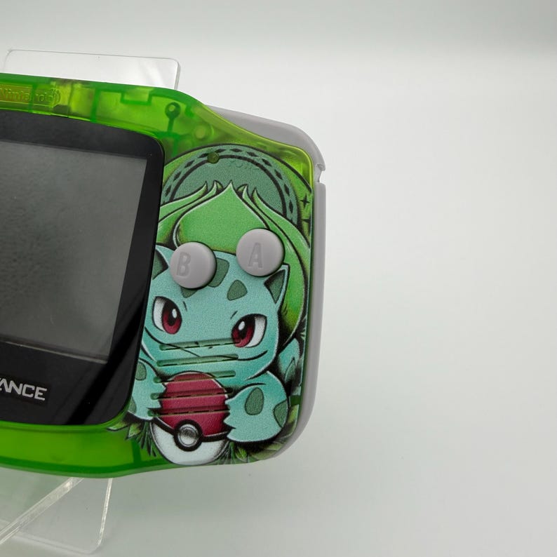 May include: A green and gray handheld gaming device with a Bulbasaur graphic. The device has a screen, gray buttons, and a green frame. The Bulbasaur graphic is in shades of green, blue, and red, holding a Pok&eacute;ball.