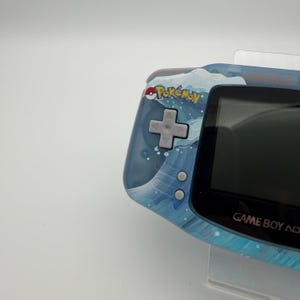 May include: A blue Game Boy Advance handheld console with a water-themed design. The console features the "Pok&eacute;mon" logo and a directional pad. The screen is black, and the buttons are gray. The console is held in a clear plastic stand.