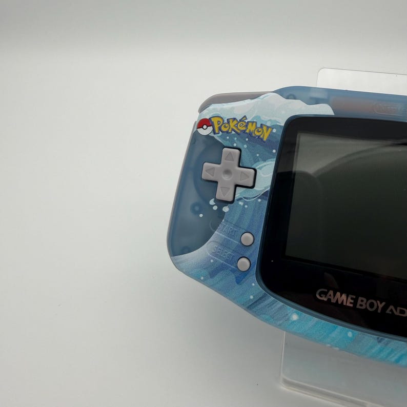 May include: A blue Game Boy Advance handheld console with a water-themed design. The console features the "Pok&eacute;mon" logo and a directional pad. The screen is black, and the buttons are gray. The console is held in a clear plastic stand.