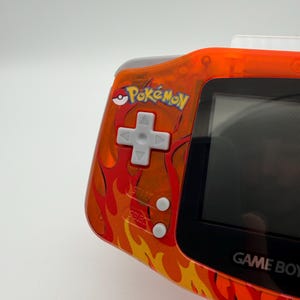 May include: Orange Game Boy Advance handheld game console with a flame design. The console features the "Pok&eacute;mon" logo and a directional pad, start, and select buttons. The screen is black.