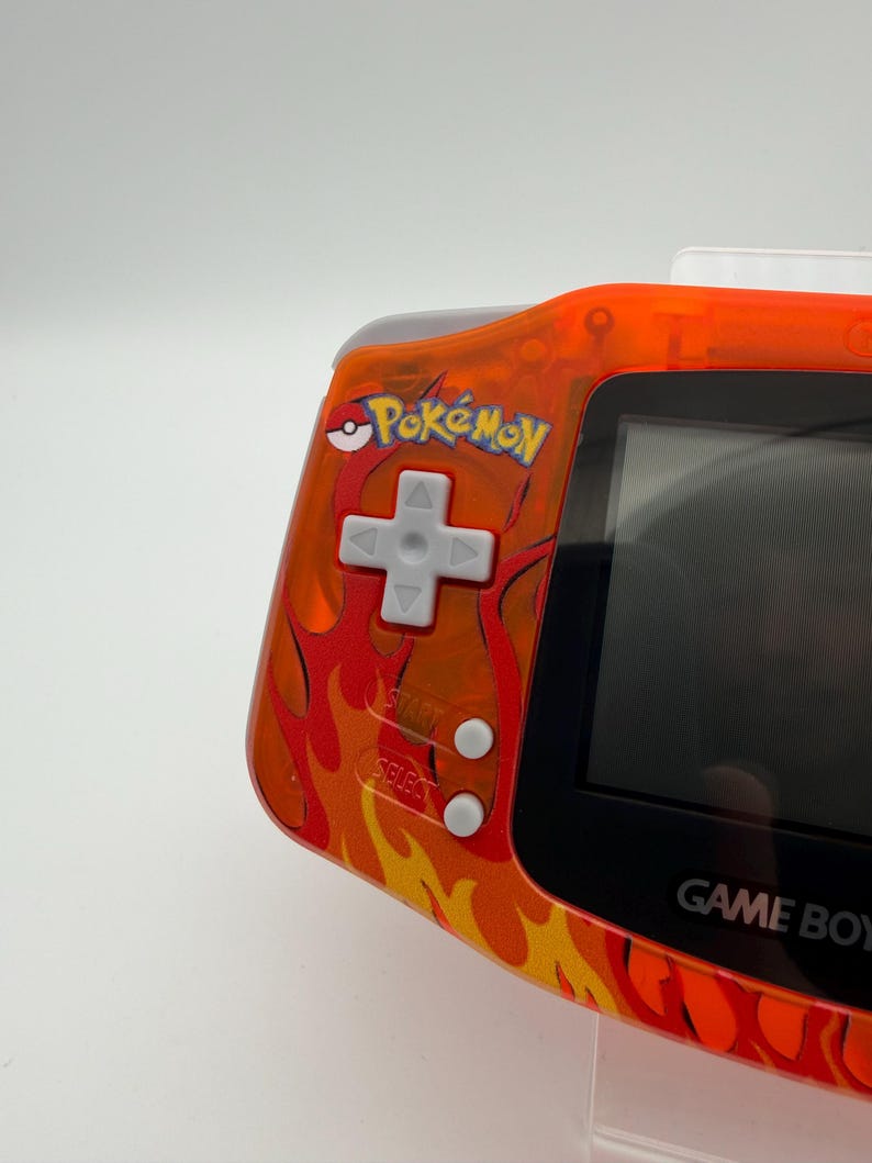 May include: Orange Game Boy Advance handheld game console with a flame design. The console features the "Pok&eacute;mon" logo and a directional pad, start, and select buttons. The screen is black.