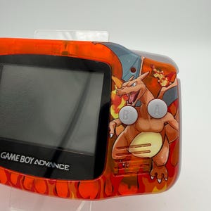 May include: An orange Game Boy Advance handheld console with a Charizard graphic. The console has a black screen and the text "GAME BOY ADVANCE" is visible. The buttons are labeled "A" and "B". The console has an orange flame design.