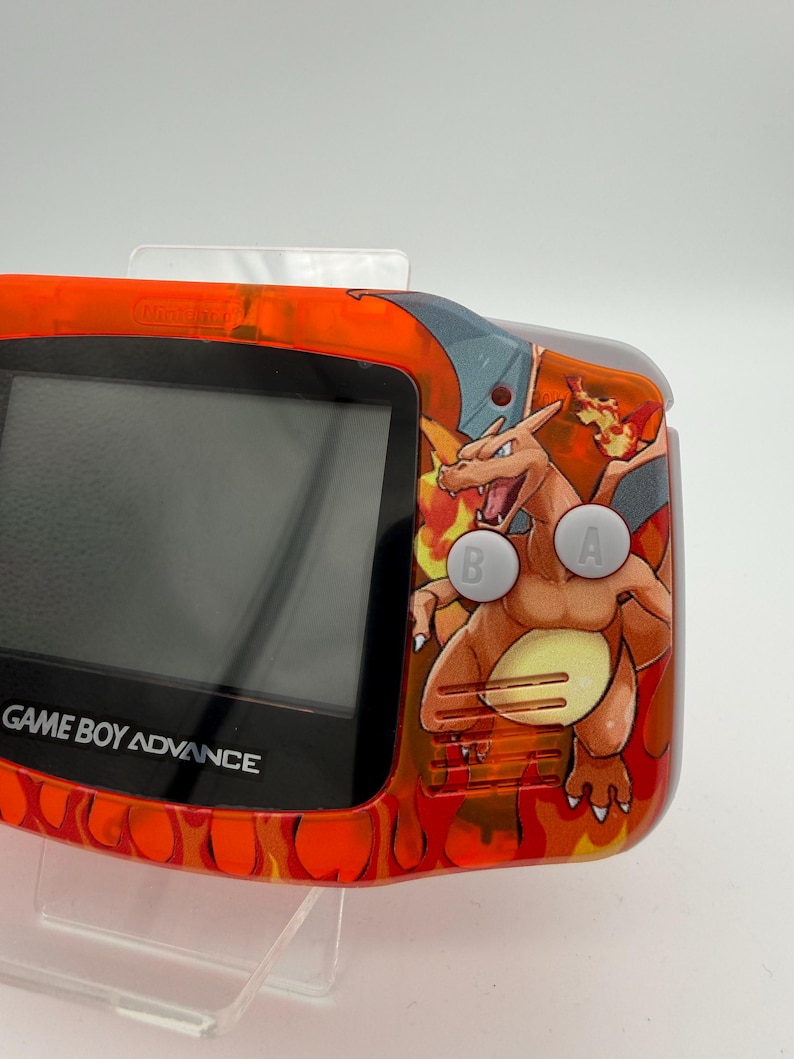 May include: An orange Game Boy Advance handheld console with a Charizard graphic. The console has a black screen and the text "GAME BOY ADVANCE" is visible. The buttons are labeled "A" and "B". The console has an orange flame design.