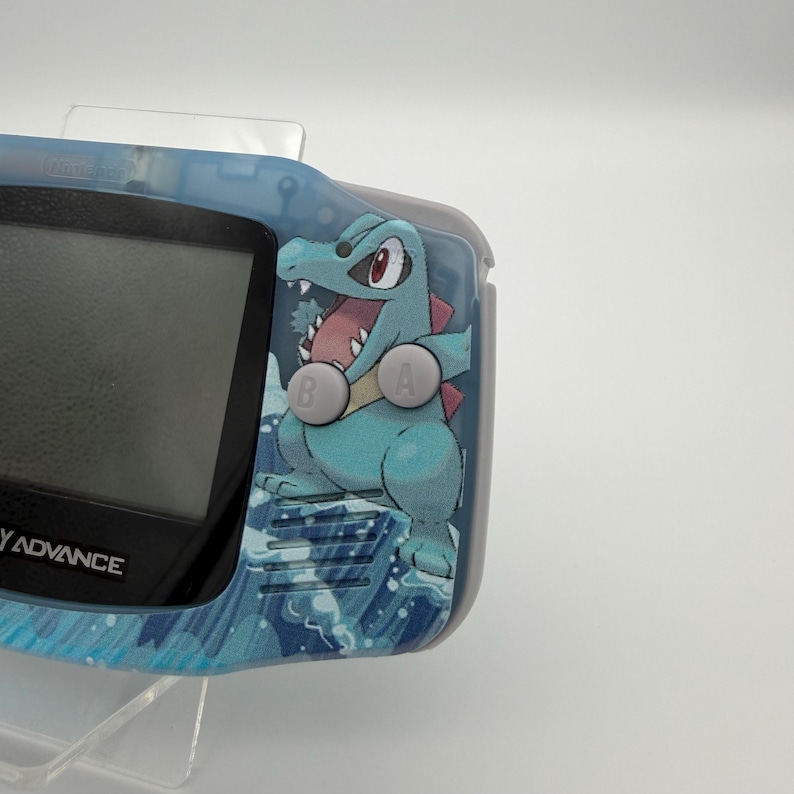 May include: A blue and gray Game Boy Advance handheld console with a custom design featuring a cartoon crocodile character. The console has a screen, buttons, and a decorative water-themed pattern.