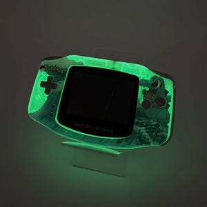 May include: A glowing green Game Boy Advance handheld console featuring a Pok&eacute;mon design. The console has a black screen and the text "GAME BOY ADVANCE". The design includes a green crocodile character and the Pok&eacute;mon logo.
