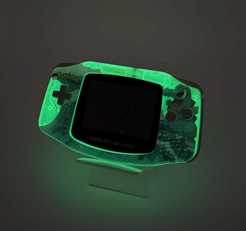 May include: A glowing green Game Boy Advance handheld console featuring a Pok&eacute;mon design. The console has a black screen and the text "GAME BOY ADVANCE". The design includes a green crocodile character and the Pok&eacute;mon logo.