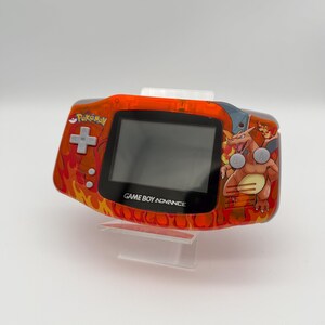 May include: An orange Game Boy Advance handheld console with a custom Pokemon design. The console features a flame pattern, the Pokemon logo, and a Charizard graphic. The text "GAME BOY ADVANCE" is visible below the screen.