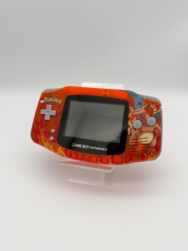 May include: An orange Game Boy Advance handheld console with a custom Pokemon design. The console features a flame pattern, the Pokemon logo, and a Charizard graphic. The text "GAME BOY ADVANCE" is visible below the screen.