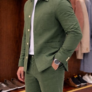 May include: Olive green suit with a matching jacket and pants. The jacket has a button-up front, a chest pocket, and two lower pockets. The suit is paired with a white t-shirt. A silver watch is visible on the wrist. The background shows a wooden display with shoes and clothing.