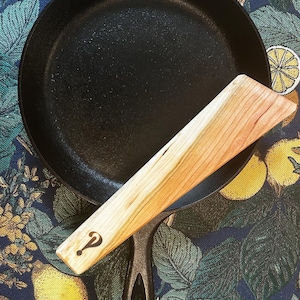May include: A black cast iron skillet with a wooden spatula. The spatula is light-colored wood with a question mark symbol. The skillet is on a blue and green floral patterned surface.