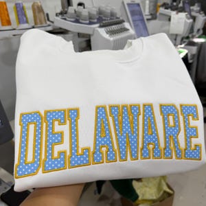 May include: A white crewneck sweatshirt featuring the word DELAWARE embroidered in large block letters. The letters are light blue with a white polka dot pattern and are outlined in a thick, vibrant yellow thread, creating a bold contrast.