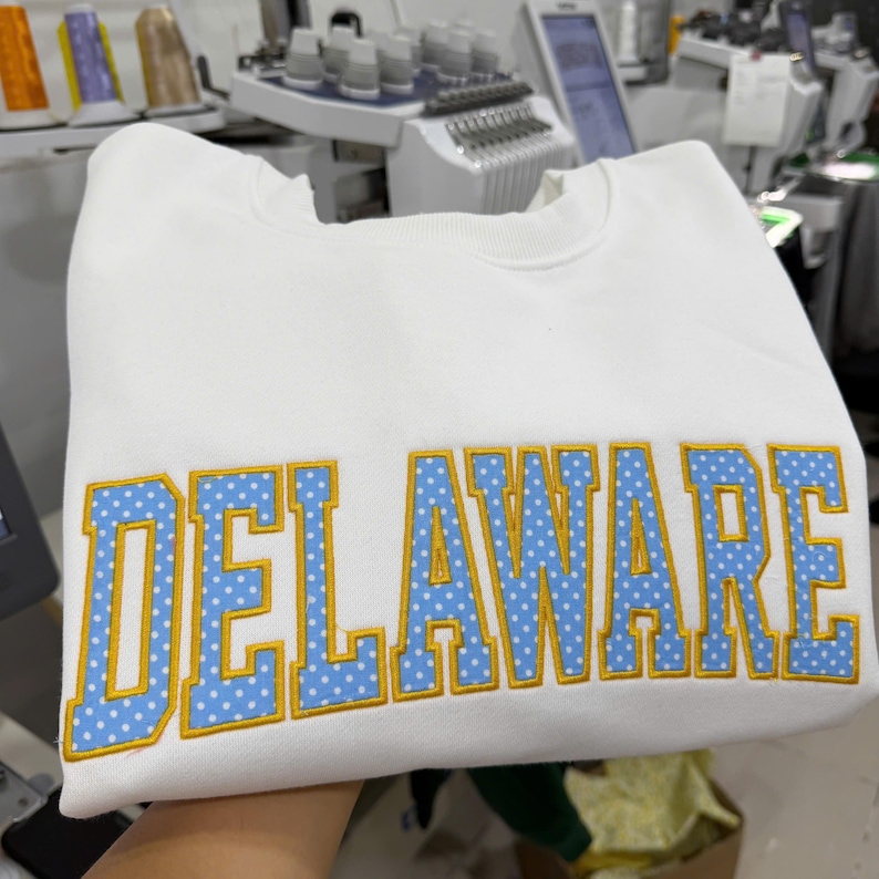 May include: A white crewneck sweatshirt featuring the word DELAWARE embroidered in large block letters. The letters are light blue with a white polka dot pattern and are outlined in a thick, vibrant yellow thread, creating a bold contrast.