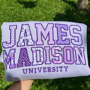 May include: A light purple sweatshirt featuring the text JAMES MADISON in large, block-style letters with various purple-toned patterns including floral, gingham, stripes, and polka dots, with the word UNIVERSITY embroidered in purple below.