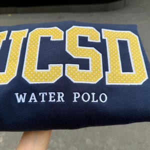 May include: A navy blue sweatshirt featuring the letters UCSD in large, yellow, white-dotted block lettering. Below the letters, the words WATER POLO are embroidered in white, capitalized, sans-serif font across the center of the garment.