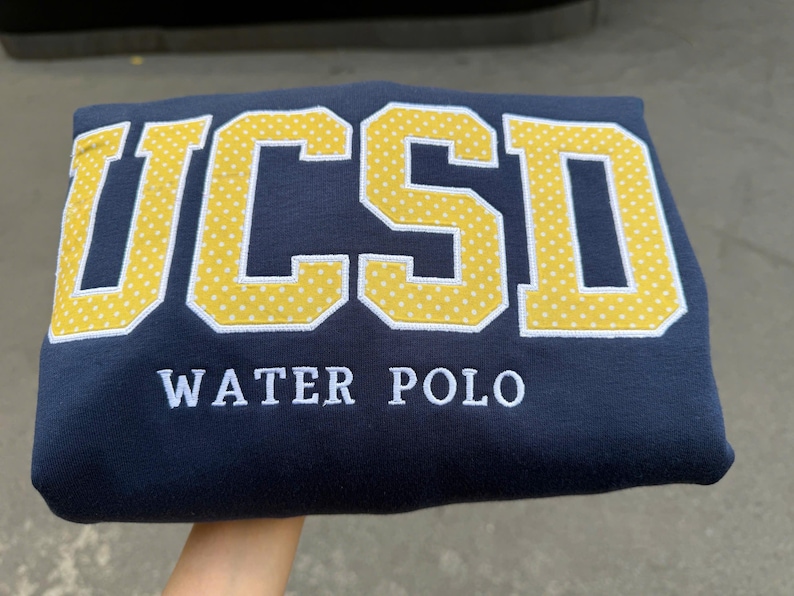 May include: A navy blue sweatshirt featuring the letters UCSD in large, yellow, white-dotted block lettering. Below the letters, the words WATER POLO are embroidered in white, capitalized, sans-serif font across the center of the garment.