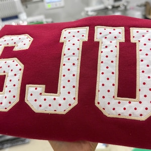 May include: A deep red sweatshirt featuring large, block-style letters that spell out SJU. The letters are crafted from white fabric with a red polka dot pattern, outlined with a light beige embroidered border that secures them to the garment.