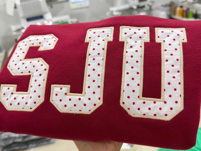 May include: A deep red sweatshirt featuring large, block-style letters that spell out SJU. The letters are crafted from white fabric with a red polka dot pattern, outlined with a light beige embroidered border that secures them to the garment.