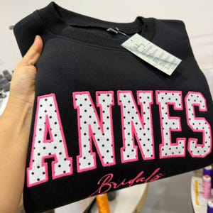 May include: A black crewneck sweatshirt featuring the word ANNES in large, white, polka-dotted block letters with bright pink outlines. Below the main text, the word Bridals is embroidered in a pink cursive script. A white tag is attached.
