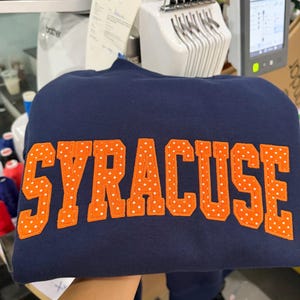 May include: A navy blue sweatshirt featuring the word SYRACUSE embroidered in large, orange block letters. Each letter is decorated with a repeating pattern of small white polka dots, creating a distinct contrast against the dark fabric.