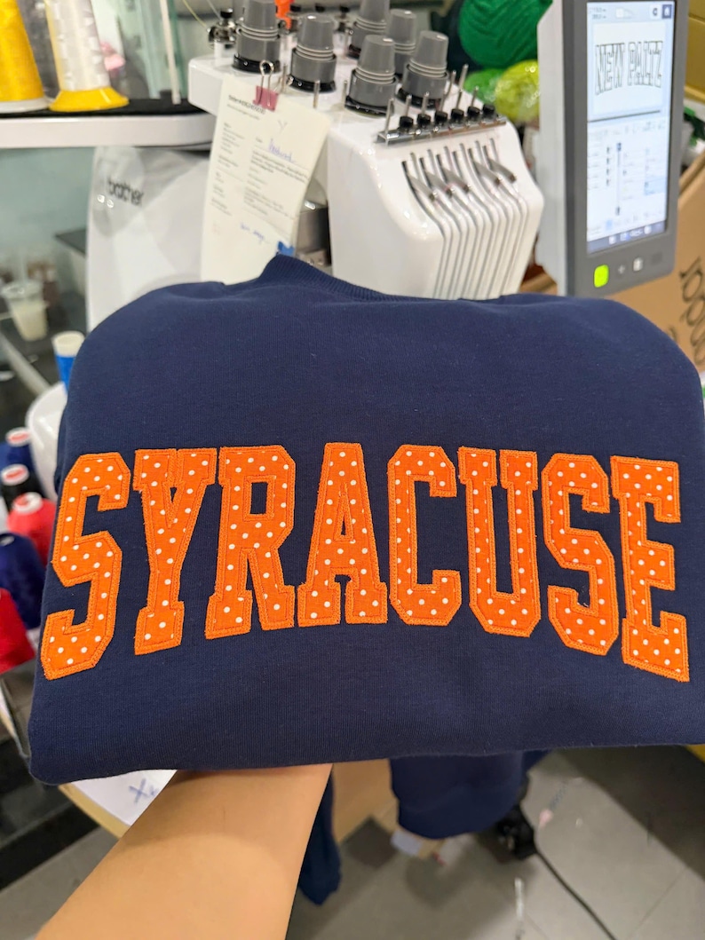 May include: A navy blue sweatshirt featuring the word SYRACUSE embroidered in large, orange block letters. Each letter is decorated with a repeating pattern of small white polka dots, creating a distinct contrast against the dark fabric.